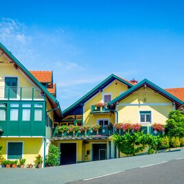 Ein malerisches Dorfhaus mit bunten Blumen und einem blauen Himmel. Die Architektur ist typisch für eine ländliche Umgebung. | © Buschenschank Tscheppe | Fam. Tscheppe