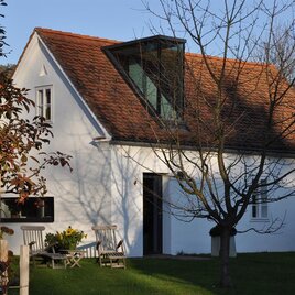 Ein modernes, weißes Haus mit einem schrägen Dach. Im Vordergrund stehen Stühle und ein Baum in einem schönen Garten.