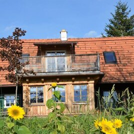 Ein charmantes Holzhaus mit einem roten Ziegeldach und einem Balkon. Im Vordergrund blühen Sonnenblumen auf einer grünen Wiese. | © Weinerlebnis Deutsch-Maier