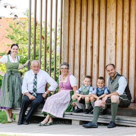 Eine fröhliche Familie sitzt auf einer Terrasse vor einer Holzwand. Alle tragen traditionelle Kleidung und lächeln in die Kamera.