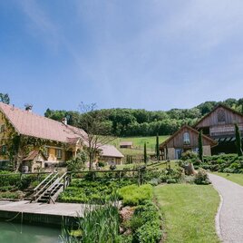 Ein malerisches Bauernhaus mit einem schönen Garten und einem kleinen Teich. Im Hintergrund sind sanfte Hügel und ein klarer Himmel zu sehen.