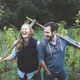 Ein fröhliches Paar in einem Weinberg, das gemeinsam lacht. Sie halten eine Schaufel und stehen zwischen Weinreben. | © Weingut  Kögl | Tamara Kögl