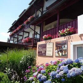 Ein charmantes Gästehaus mit blühenden Balkonpflanzen und einer einladenden Fassade. Der Garten ist üppig mit blauen Hydrangeas und grünen Pflanzen gestaltet. | © Weststeirischer Hof