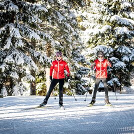 Zwei Skifahrer in roten Jacken stehen auf einer schneebedeckten Loipe. Um sie herum sind hohe, verschneite Bäume. | © Orthofer