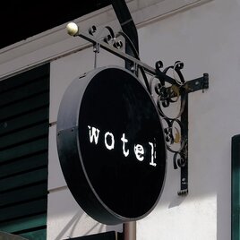 Ein runder, schwarzer Schild mit dem Schriftzug "wotel" hängt an einer Wand. Es ist elegant und schlicht gestaltet.