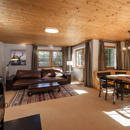 Ein gemütlicher Wohnbereich mit einem Sofa, einem Esstisch und großen Fenstern. Holzdecke und warme Farben schaffen eine einladende Atmosphäre.