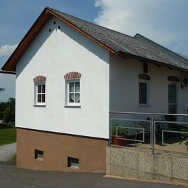 Ein modernes, helles Haus mit einem gemütlichen Balkon. Im Hintergrund sind grüne Bäume und eine gepflegte Landschaft zu sehen.